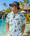 A man wearing a light blue Hawaiian shirt with a floral print and a navy blue bucket hat with 'Hawaii' printed on the front is standing by a tropical resort pool with palm trees in the background.