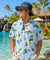 A man wearing a light blue Hawaiian shirt with a floral print and a navy blue bucket hat with 'Hawaii' printed on the front is standing by a tropical resort pool with palm trees in the background.