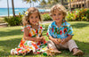 Children in colorful Hawaiian outfits sitting on grass with a beach and palm trees in the background